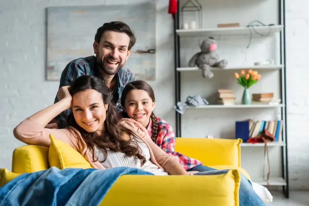  Melhores bairros de São Paulo para viver ou investir: imagem de pai, mãe e filha sorrindo em uma sala cinza, com sofá amarelo e armário com livros, flores e ursinho