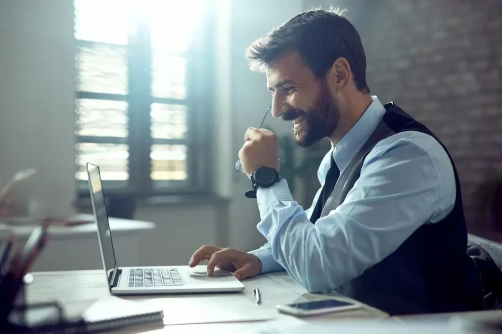Investir em Franquia Imobiliária: homem sentado na mesa e sorrindo na frente de um notebook, perto de janelas e paredes