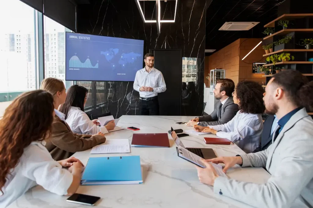 Investir em franquia imobiliária: equipe composta de 3 homens e 4 mulheres discutindo resultados da empresa em uma mesa. Um homem está de pé apresentando gráficos.