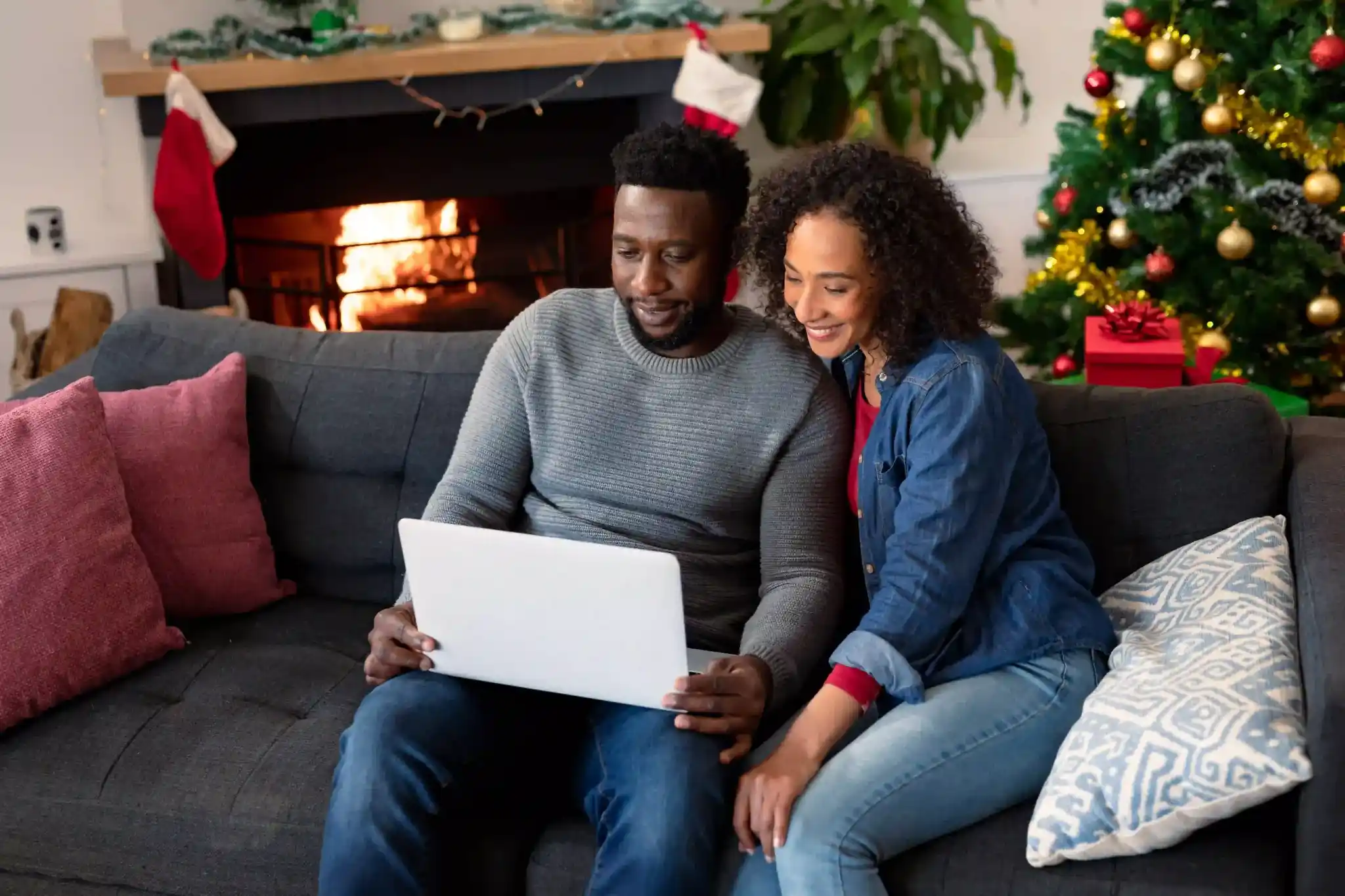 Decorações de Natal: casal sentado no sofá olhando notebook com árvore de natal e decorações natalinas de fundo