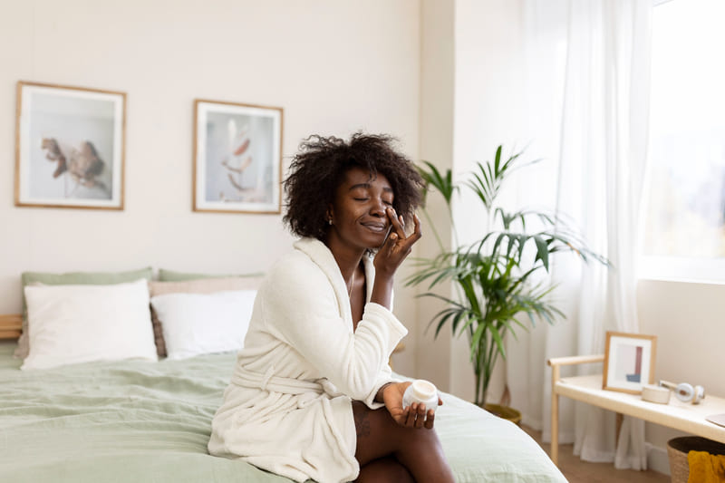 Mercado imobiliário: mulher sentada na cama do seu quarto enquanto passa produto no rosto - Auxiliadora Predial