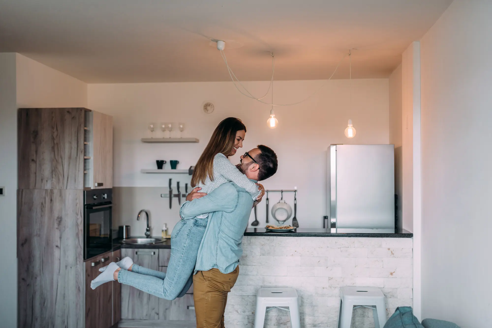 5 Melhores tipos de imóveis para viver a dois: imagem de casal se abraçando perto da cozinha, com fogão, geladeira, cadeiras e acessórios.