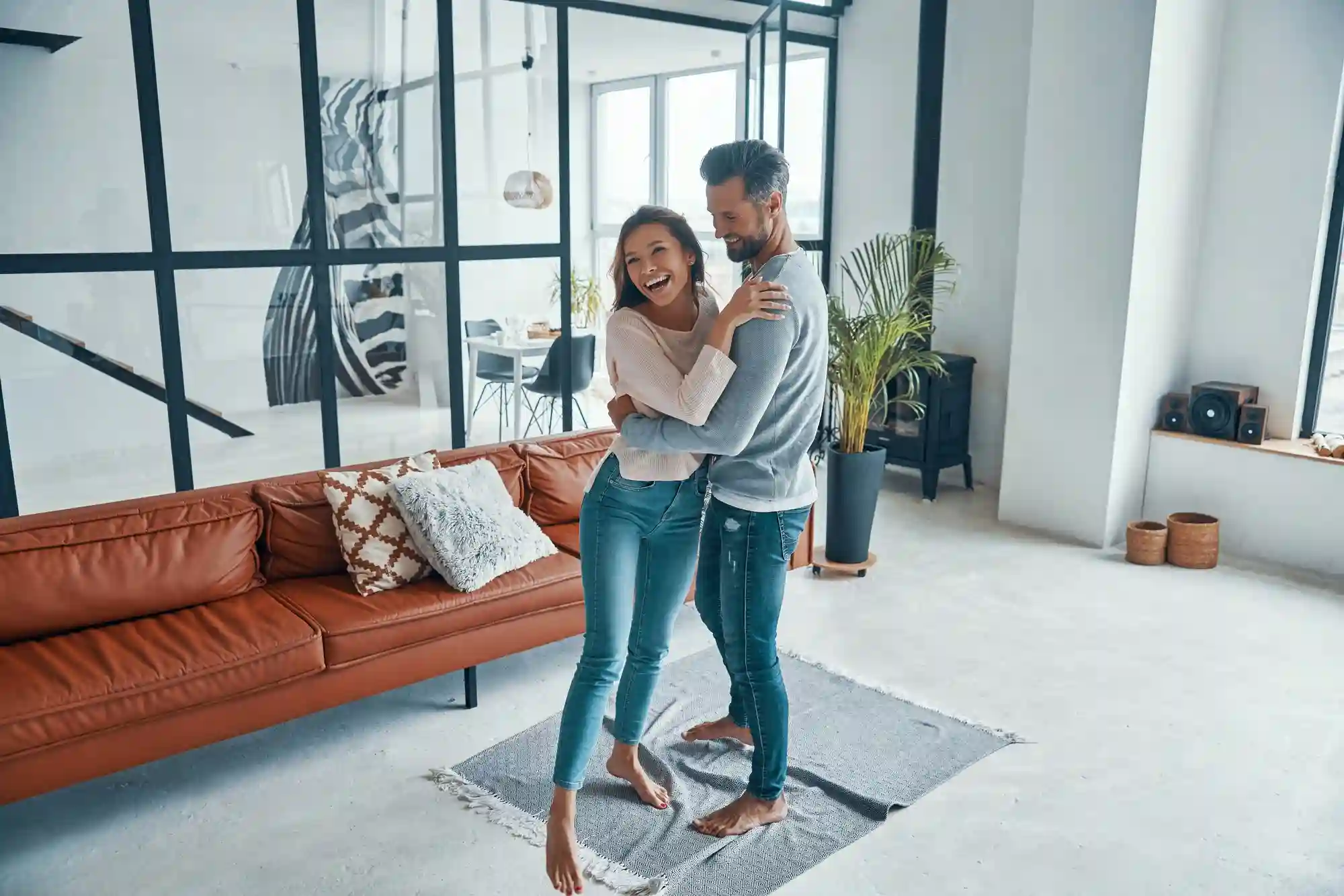 5 Melhores tipos de imóveis para viver a dois: casal se abraçando em seu flat, no centro da sala e em cima de um tapete. Na foto vemos sofá, plantas, janelas e varanda.
